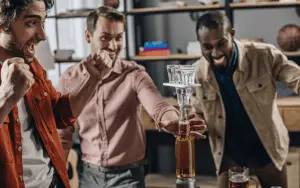 Eine lebhafte Gruppe von Freunden, lachend und unterhaltsam, während sie auf einer aufregenden Pub Crawl Tour durch die belebten Straßen einer Stadt unterwegs sind. Die Gruppe ist in verschiedenen Bars und Pubs und Clubs zu sehen, wo sie in fröhlicher Stimmung verschiedene Getränke probieren und die einzigartige Atmosphäre jeder Location genießen. Die bunten Lichter der Bars leuchten in der Nacht, und die Teilnehmer tauschen fröhliche Gespräche und Toasts aus, während sie die lokale Bar-Szene erkunden. Ein unvergessliches Abenteuer, das Freundschaften schmiedet und die Nacht in lebendige Erinnerungen verwandelt. Junggesellenabschied | JGA