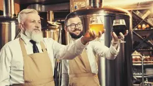 Zwei Männer halten jeweils ein Bierglas vor mehreren Braukesseln in die Luft